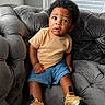 child, toddler, baby, portrait, sitting, sofa, couch, curly_hair, smiling, wide_eyes, indoor, living_room, window, blinds, gray_furniture, tan_shirt, denim_shorts, golden_shoes, fashion, photography