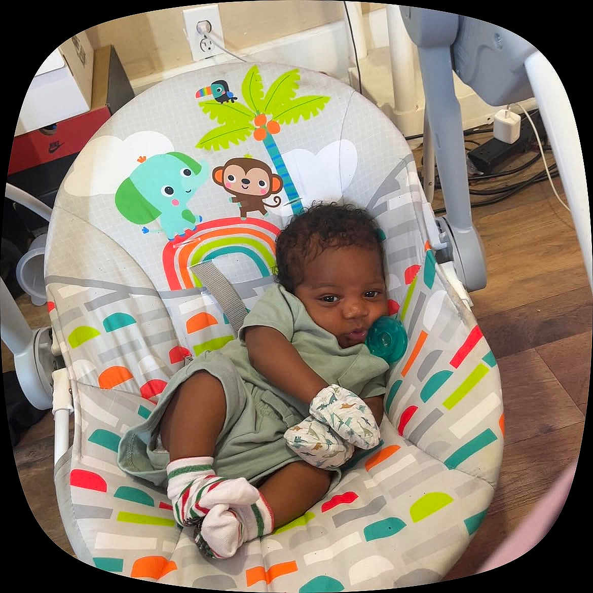 Nehemiah Jr is registered to the contest to win money with this photo: baby, infant, baby_seat, mittens, socks, pacifier, wooden_floor, colorful, animal_illustration, monkey, elephant, rainbow, palm_tree, indoors, child, cute, relaxed, clothing, floor, seat
