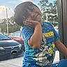 Zealand is registered to the contest to win money with this photo: child, portrait, smiling, happy, playful, posing, hand_on_chin, short_hair, blue_shirt, cartoon_shirt, blue_pants, window, indoors, outdoor_view, parked_cars, street, traffic_lights, trees, daylight, colorful
