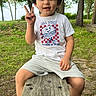 child, boy, smiling, peace_sign, outdoors, park, trees, nature, lake, wooden_bench, sitting, portrait, short_hair, casual_clothing, t_shirt, shorts, sandals, sunlight, grass, happy