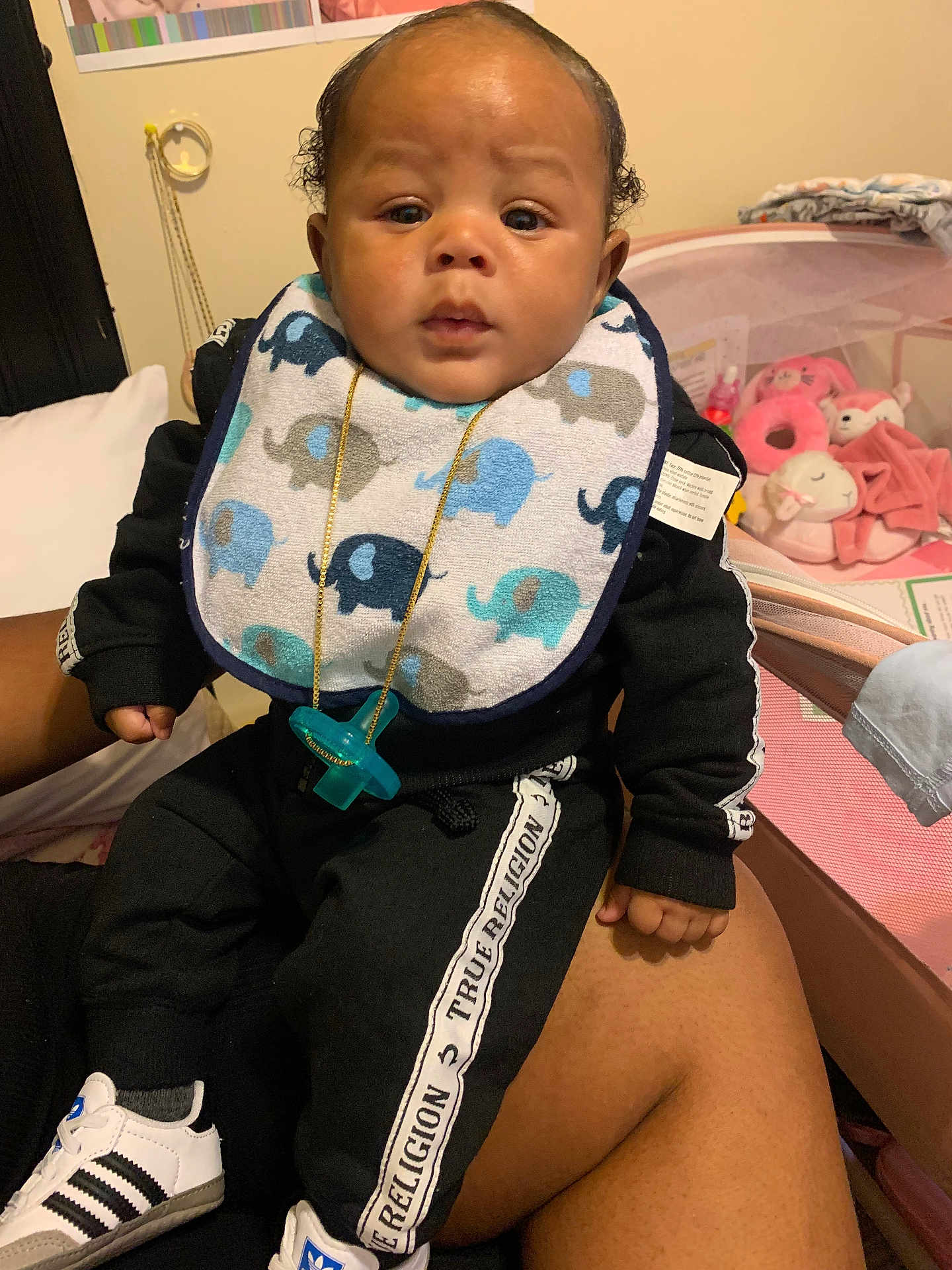 Jyzere is registered to the contest to win money with this photo: baby, infant, bib, pacifier, tracksuit, black_clothing, elephant_pattern, sneakers, adults_lap, plush_toys, nursery, indoor, child, cute, person, footwear, toy, seated, portrait, home