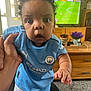 baby, child, indoor, football_kit, manchester_city, sportswear, television, soccer, hand, curly_hair, person, carpet, furniture, plant, flower_pot, living_room, sports, cute, wide_eyes, curious