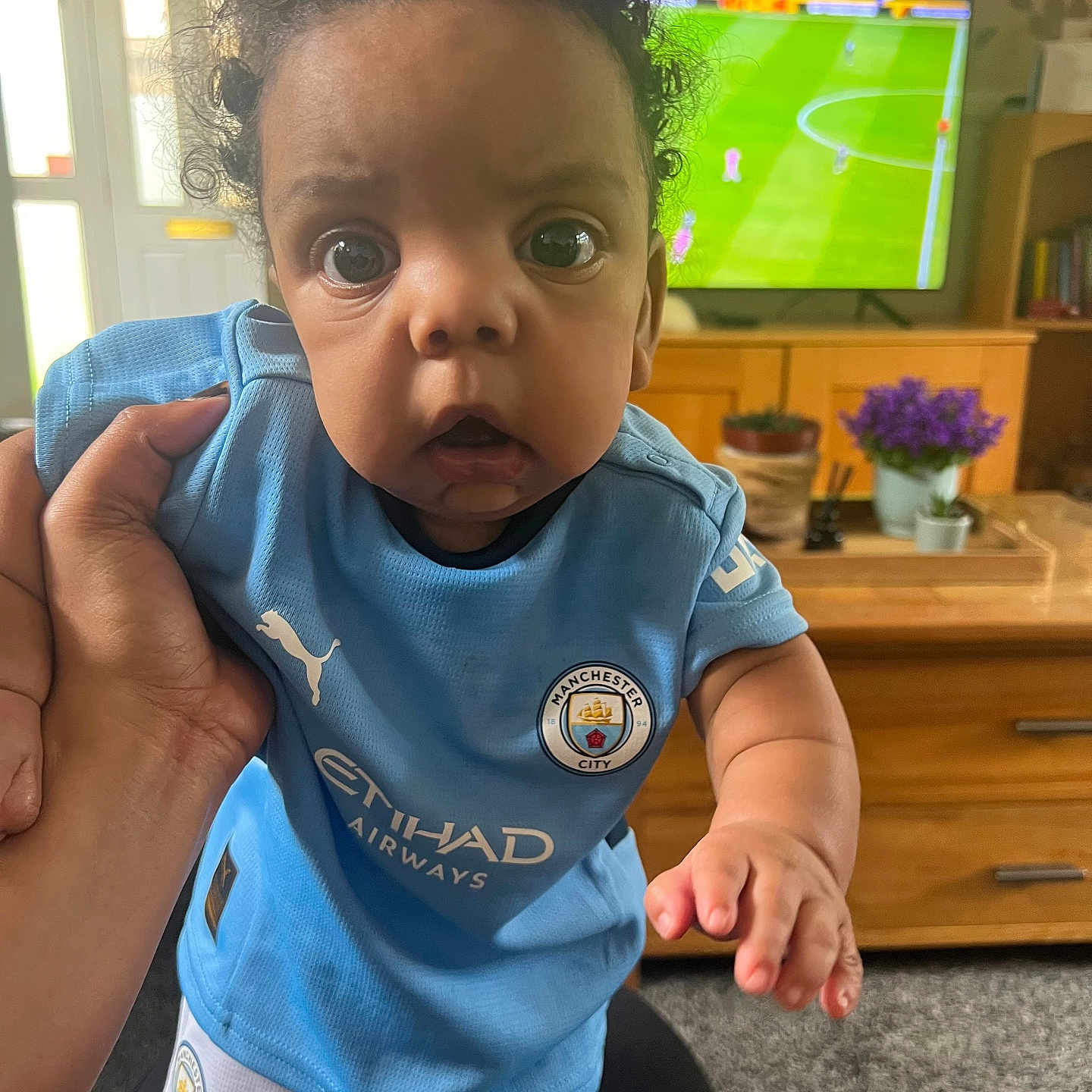 Suliman joined the competition — help win amazing prizes! baby, carpet, child, curious, curly_hair, cute, flower_pot, football_kit, furniture, hand, indoor, living_room, manchester_city, person, plant, soccer, sports, sportswear, television, wide_eyes