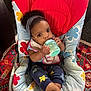 baby, child, bottle, feeding, chair, headband, curly_hair, clothing, bib, feet, indoor, carpet, pattern, colorful, cute, person, infant, portrait, sitting, hand