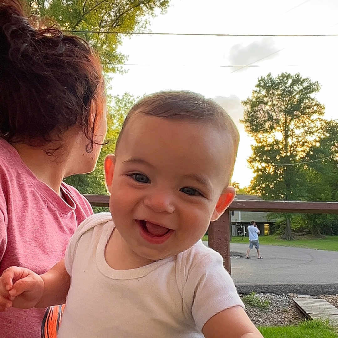 Bentley is registered to the contest to win money with this photo: baby, casual_clothing, child, curly_hair, daylight, fringed_canopy, glass_table, greenery, happy, nature, outdoor, patio, person, reaching_hand, smiling, sunset, table, trees, white_shirt, woman