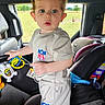 Charlie is registered to the contest to win money with this photo: toddler, child, curly_hair, blue_eyes, clothing, tshirt, shorts, toy, car_seat, vehicle_interior, hand, face, person, pointing, window, outdoor_view, seatbelt, blanket, greenery, park