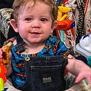 Charlie is registered to the contest to win money with this photo: toddler, child, baby, smiling, curly_hair, denim_overalls, blue_shirt, toy, colorful, indoor, face, expression, happy, snack, play_area, patterned_blanket, furniture, closeup, messy_face, person