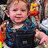 toddler, child, baby, smiling, curly_hair, denim_overalls, blue_shirt, toy, colorful, indoor, face, expression, happy, snack, play_area, patterned_blanket, furniture, closeup, messy_face, person