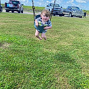 Tobias is registered to the contest to win money with this photo: child, toddler, swing, playground, grass, sky, clouds, pickup_truck, vehicle, baby_swing, smiling, outdoor, summer, park, swing_chain, bucket_seat, barefoot, shirt, face, parking_lot