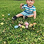 child, toddler, boy, striped_clothing, blue_stripes, grass, easter_eggs, camouflage_egg, clover, sitting, outdoor, portrait, blond_hair, black_shoes, toy, truck_wheel, vehicle, lawn, curious_expression, greenery