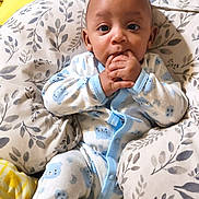 Chosen is registered to the contest to win money with this photo: baby, infant, child, portrait, indoors, bed, pillow, blanket, pajamas, blue_clothing, yellow_blanket, sitting, lying_down, wide_eyed, hand_to_mouth, smiling, cozy, bedding, floral_pattern, home