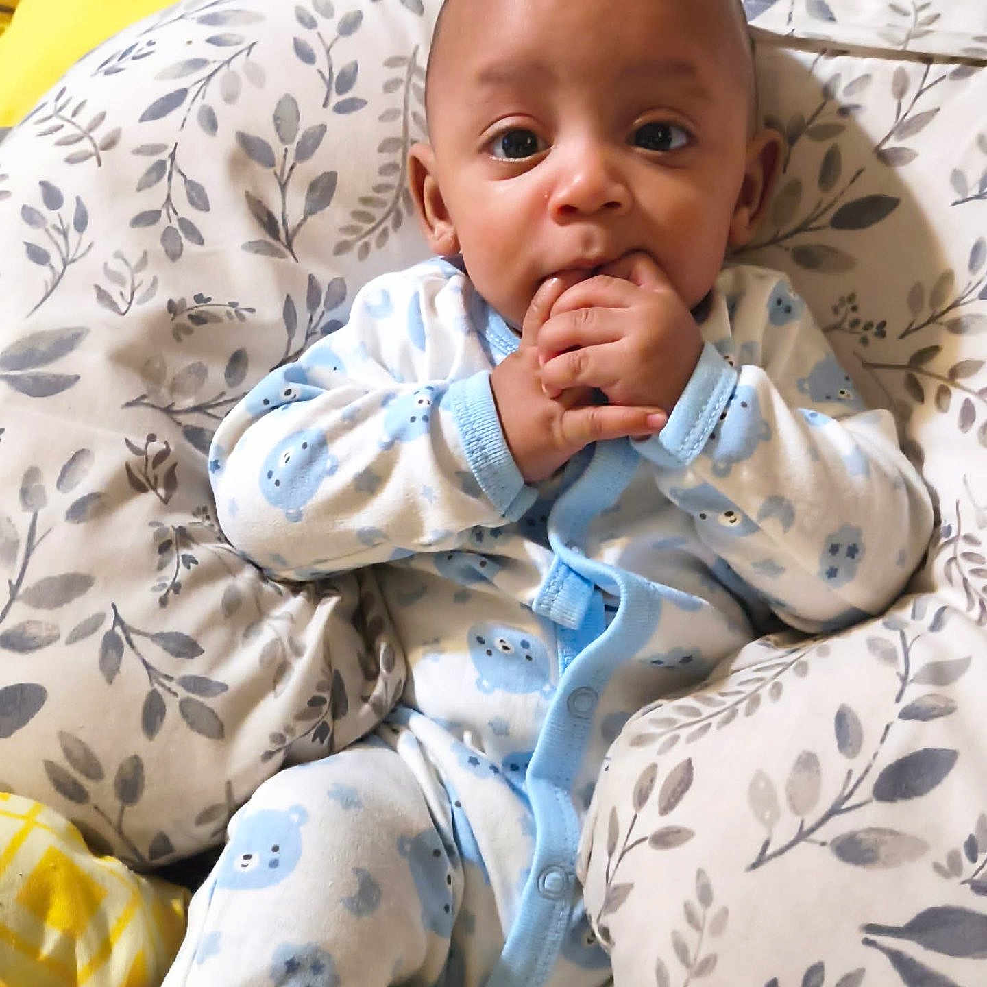 Chosen is registered to the contest to win money with this photo: baby, infant, child, portrait, indoors, bed, pillow, blanket, pajamas, blue_clothing, yellow_blanket, sitting, lying_down, wide_eyed, hand_to_mouth, smiling, cozy, bedding, floral_pattern, home