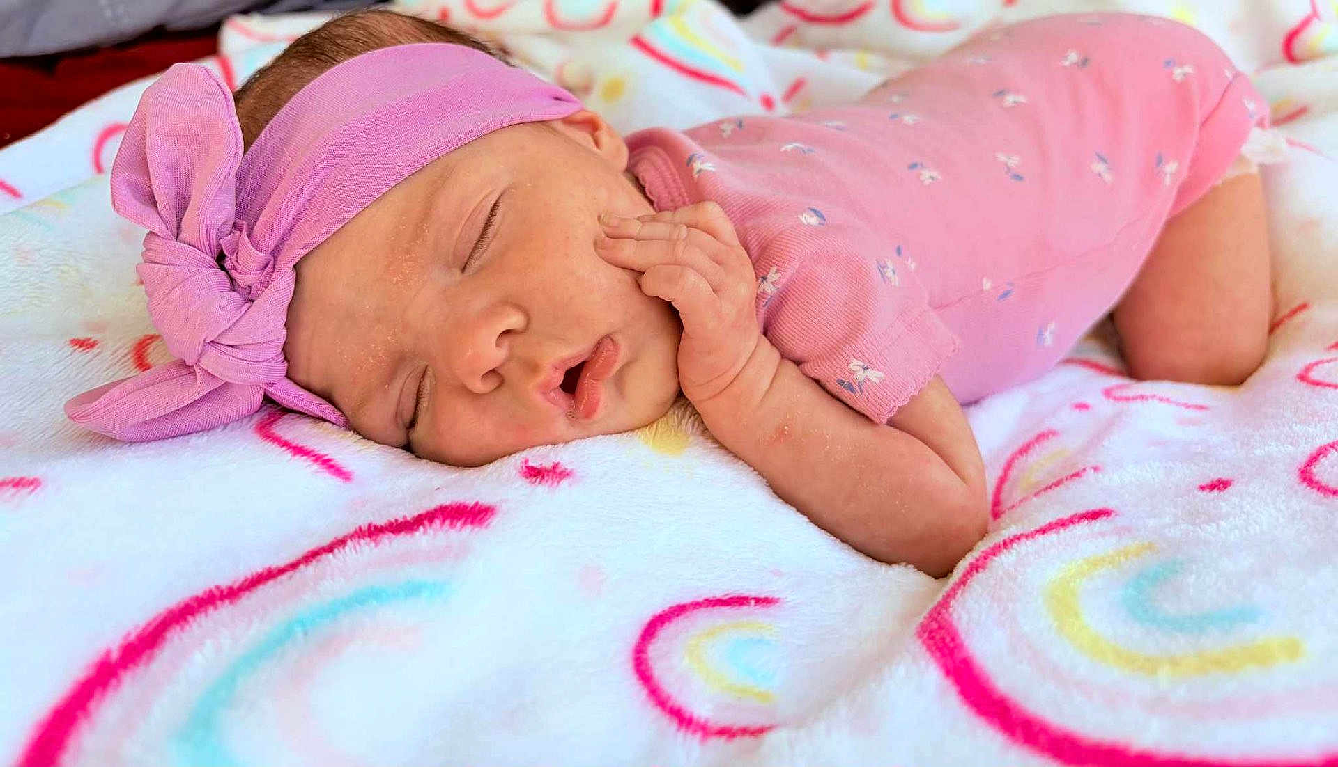 Rylee is registered to the contest to win money with this photo: baby, sleeping, pink_clothing, headband, blanket, rainbow_pattern, infant, cute, soft_texture, resting, child, peaceful, newborn, indoors, portrait, hand, face, cozy, comfort, closeup