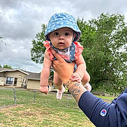 Melanie is registered to the contest to win money with this photo: baby, child, hat, blue_hat, pink_clothing, tattoo, arm, hand, outdoor, cloudy_sky, grass, fence, house, tree, person, cute, portrait, summer, holding, expression