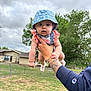 baby, child, hat, blue_hat, pink_clothing, tattoo, arm, hand, outdoor, cloudy_sky, grass, fence, house, tree, person, cute, portrait, summer, holding, expression