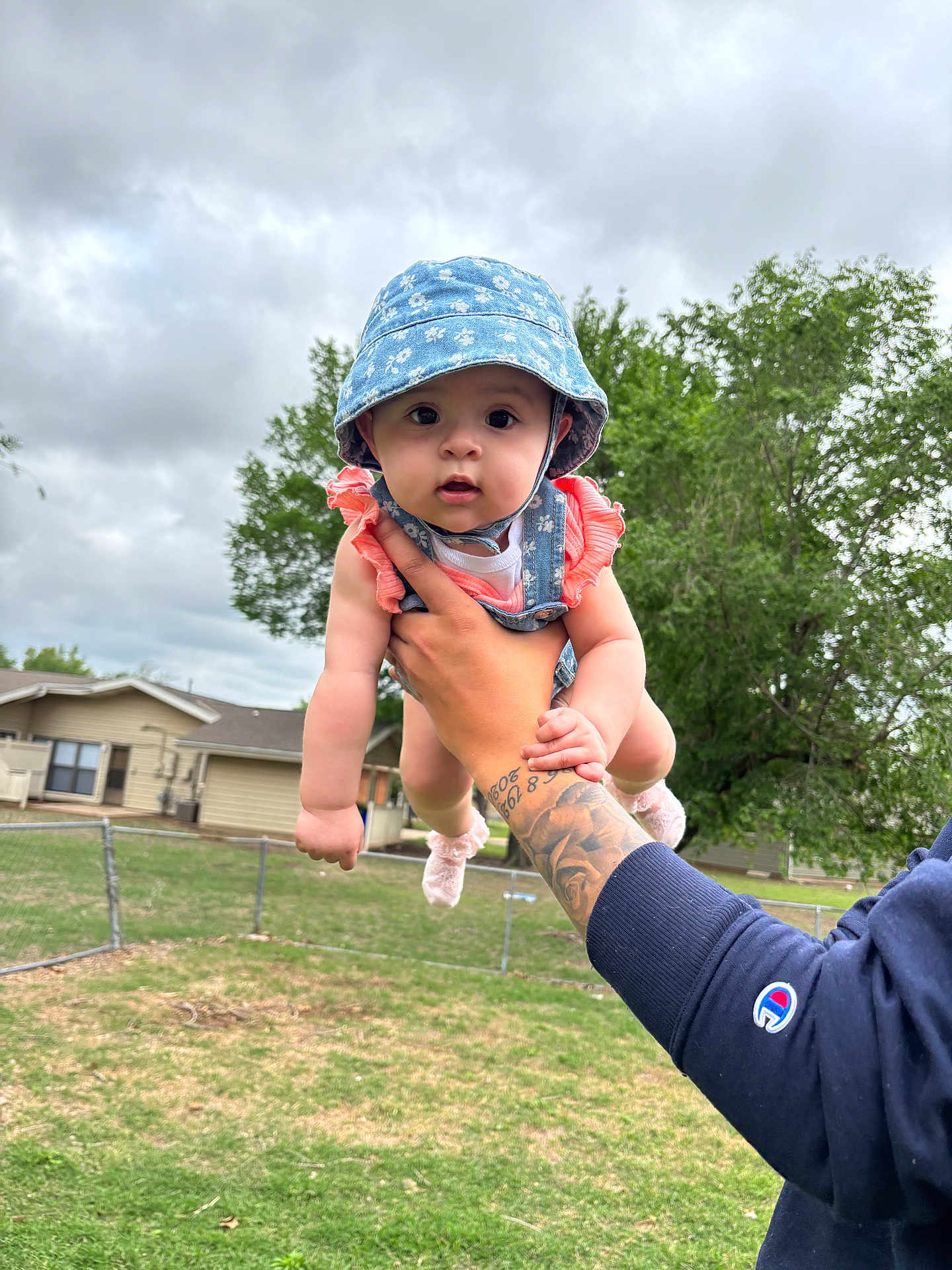 Melanie is registered to the contest to win money with this photo: baby, child, hat, blue_hat, pink_clothing, tattoo, arm, hand, outdoor, cloudy_sky, grass, fence, house, tree, person, cute, portrait, summer, holding, expression