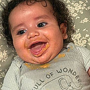 Alijah is registered to the contest to win money with this photo: baby, child, curly_hair, smiling, happy, food_mess, face, clothing, shirt, elephant_graphic, text_on_clothing, blanket, floral_pattern, lying_down, infant, cute, joyful, portrait, close_up, messy