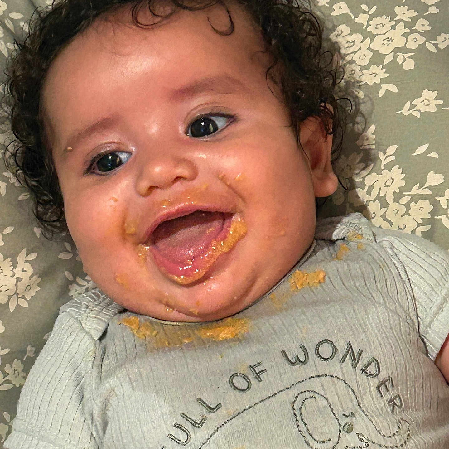 Alijah is registered to the contest to win money with this photo: baby, child, curly_hair, smiling, happy, food_mess, face, clothing, shirt, elephant_graphic, text_on_clothing, blanket, floral_pattern, lying_down, infant, cute, joyful, portrait, close_up, messy