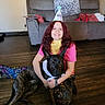 bandana, black_dog, blanket, casual_clothing, couch, cushion, dog, floor, happy, home, hugging, indoor, living_room, party_hat, person, pet, red_hair, seated, smiling, wooden_floor
