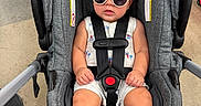 Raidel is registered to the contest to win money with this photo: baby, child, sunglasses, car_seat, gray, infant, sitting, footwear, sandals, clothing, safety_harness, cute, indoor, floor, person, fashion, accessory, legs, head, summer