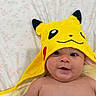 baby, child, pikachu, yellow, hooded_towel, face, skin, smile, eyes, blanket, floral_pattern, indoors, cute, infant, portrait, person, childhood, soft_texture, happy, lying_down