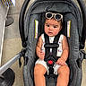 baby, car_seat, sunglasses, child, infant, footwear, straps, concrete_floor, shopping_area, person, clothing, summer_wear, foot, red_bracelet, indoor, seat, expression, cute, toddler, safety_harness