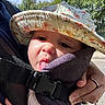 baby, child, hat, floral, tongue, face, outdoor, sunlight, trees, carrier, hand, ring, person, clothing, nature, expression, cute, infant, closeup, daytime