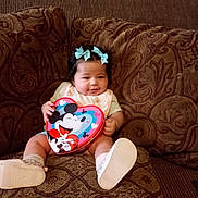 Rosie joined the competition — help win amazing prizes! baby, child, cute, mickey_mouse, heart_shaped_box, blue_bows, couch, patterned_fabric, smiling, sitting, footwear, indoor, portrait, infant, toy, holding, happy, person, decor, home