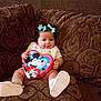 baby, child, cute, mickey_mouse, heart_shaped_box, blue_bows, couch, patterned_fabric, smiling, sitting, footwear, indoor, portrait, infant, toy, holding, happy, person, decor, home