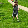toddler, child, grass, lawn, sidewalk, toy_wheelbarrow, camouflage_hat, tank_top, shorts, rain_boots, greenery, outdoor, daylight, smiling, walking, vehicles, toy, play, happy, nature