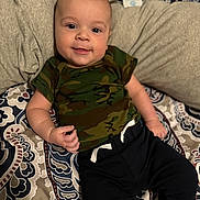 Aiden joined the competition — help win amazing prizes! baby, infant, smile, camo_shirt, black_pants, patterned_bedspread, lying_down, happy, cute, child, person, indoors, soft_lighting, casual_clothing, portrait, young_child, socks, cozy, relaxed, face