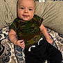 baby, infant, smile, camo_shirt, black_pants, patterned_bedspread, lying_down, happy, cute, child, person, indoors, soft_lighting, casual_clothing, portrait, young_child, socks, cozy, relaxed, face