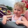 amusement_park, background, child, cloudy_sky, colorful, curly_hair, daytime, entertainment, face, family, fun, hand, nail_polish, outdoor, park, people, pink_tank_top, ride, smile, toddler