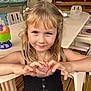 child, girl, blonde_hair, blue_eyes, smiling, hand_gesture, heart_shape, black_dress, plastic_chair, table, toy, indoor, wooden_floor, casual, portrait, cute, happy, person, young, summer