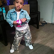 Gianna is registered to the contest to win money with this photo: toddler, child, denim_jacket, camouflage_pants, indoor, person, standing, floor, furniture, plastic_bottle, box, hair_bun, white_shoes, casual_clothing, young_child, holding_object, home_interior, expression, flooring, shadow