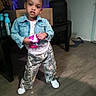 toddler, child, denim_jacket, camouflage_pants, indoor, person, standing, floor, furniture, plastic_bottle, box, hair_bun, white_shoes, casual_clothing, young_child, holding_object, home_interior, expression, flooring, shadow