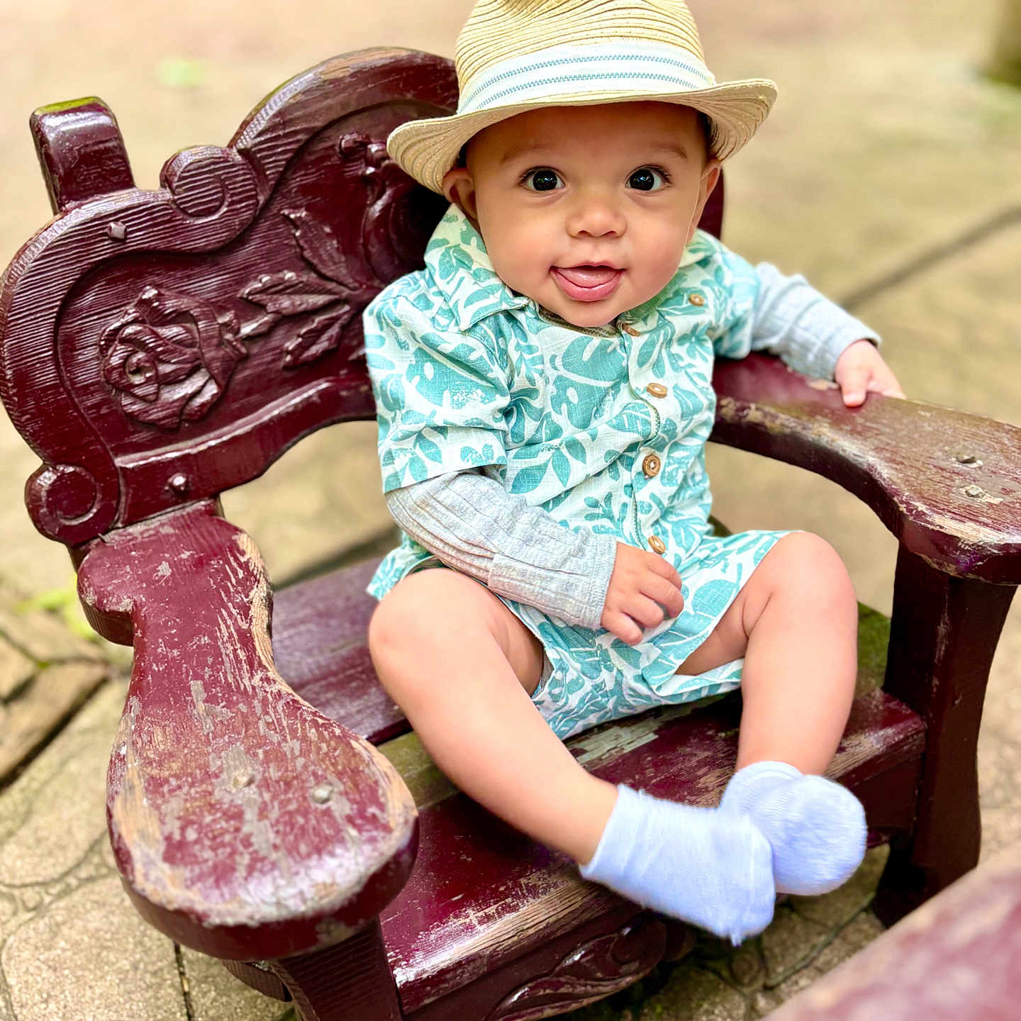 Jasper is registered to the contest to win money with this photo: baby, child, hat, straw_hat, smile, tongue_out, wooden_chair, outdoor, greenery, patterned_clothing, happy, cute, sitting, portrait, infant, natural_light, eyes, face, footwear, sock