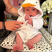 Sonny joined the competition — help win amazing prizes! baby, child, person, white_robe, duck_hood, orange_beak, sitting, hands, adult_hand, nail_polish, cushion, polka_dots, table, flowers, vase, decor, pink_wall, heart_decorations, indoor, portrait