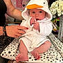 baby, child, person, white_robe, duck_hood, orange_beak, sitting, hands, adult_hand, nail_polish, cushion, polka_dots, table, flowers, vase, decor, pink_wall, heart_decorations, indoor, portrait