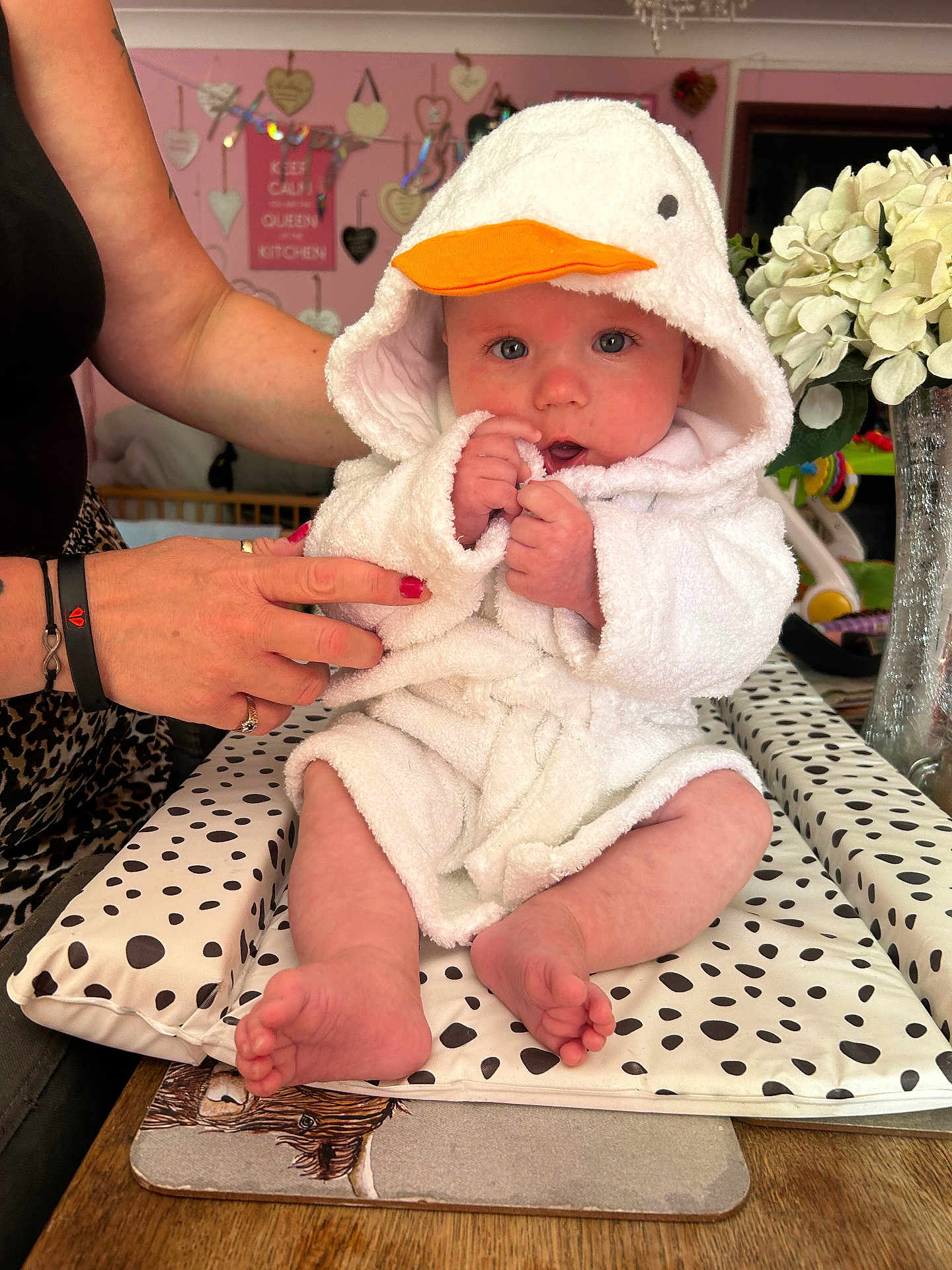 Sonny joined the competition — help win amazing prizes! baby, child, person, white_robe, duck_hood, orange_beak, sitting, hands, adult_hand, nail_polish, cushion, polka_dots, table, flowers, vase, decor, pink_wall, heart_decorations, indoor, portrait