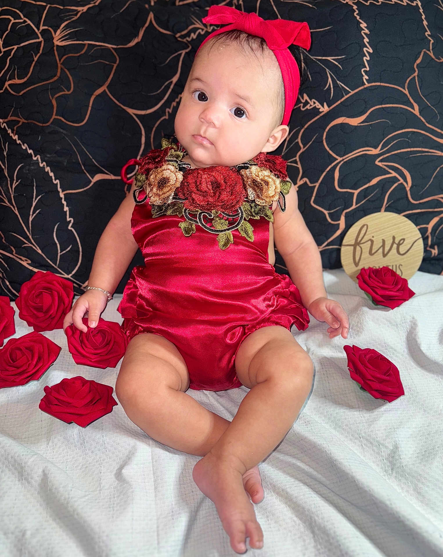 Ileana is registered to the contest to win money with this photo: baby, child, red_outfit, headband, roses, artificial_flowers, white_blanket, black_background, floral_pattern, wooden_sign, sitting, portrait, cute, celebration, indoor, person, young_child, decor, fabric, accessory
