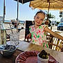 child, girl, smiling, outdoor, cafe, table, food, plate, sausage, pickles, glass, water, umbrella, beach, bicycle, people, sunny, daytime, dress, portrait