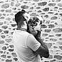 man, baby, child, holding, kissing, laughing, black_and_white, stone_wall, affection, joy, outdoor, casual_clothing, short_sleeves, portrait, parent_and_child, emotion, love, smile, family, tenderness