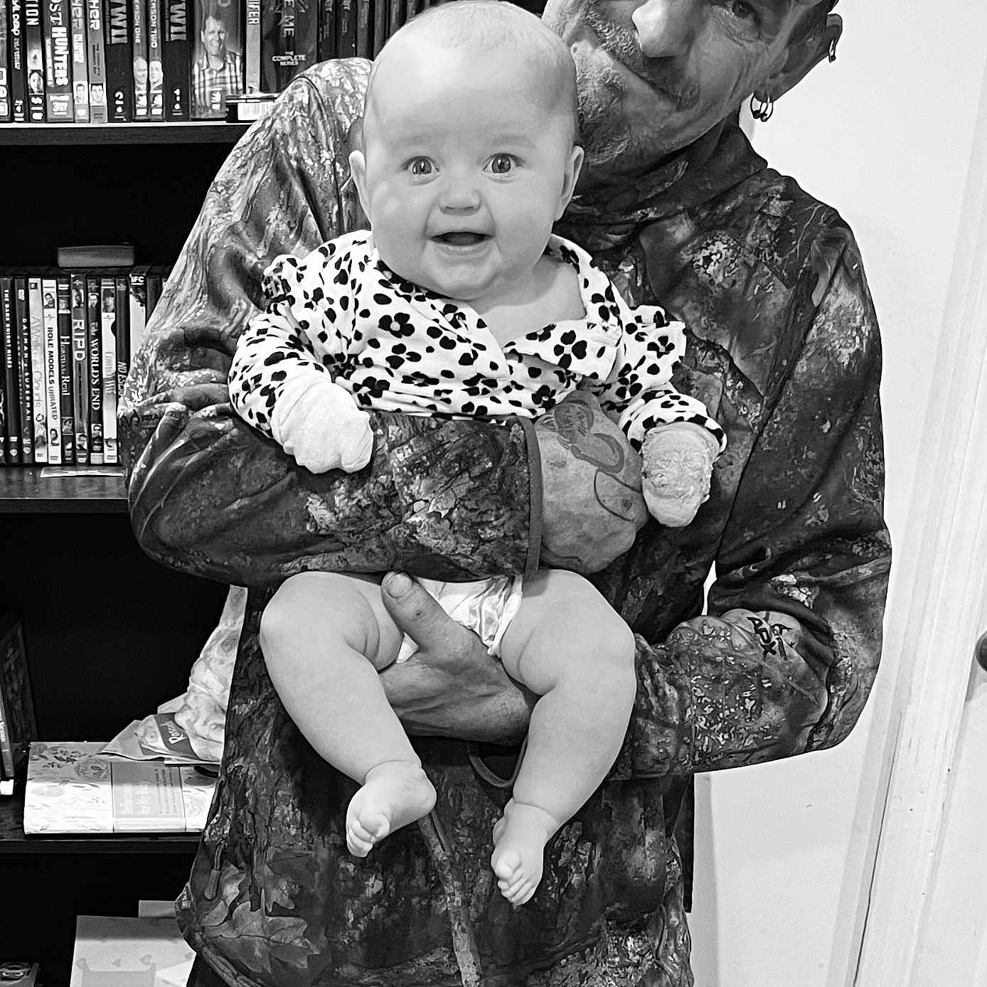 Alena is registered to the contest to win money with this photo: baby, black_and_white, bookshelf, camo_hoodie, cap, child, clothing, dvds, earrings, expression, face, happy, holding, indoor, infant, man, person, portrait, smile, tattoo