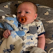 Easton is registered to the contest to win money with this photo: baby, pacifier, stuffed_animal, blanket, cushion, patterned, clothing, infant, cute, soft_toy, blue, white, face, head, lying_down, comfort, sleepwear, child, indoors, resting