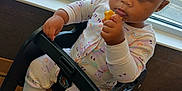 Icslyn is registered to the contest to win money with this photo: baby, child, high_chair, pajamas, snack, indoor, window, floor_tiles, curly_hair, expression, person, seat, pink_socks, food, hand, face, looking, young_child, cute, furniture