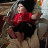 baby, beanie, red_shirt, milk_carton_design, baby_seat, blanket, carpet, indoor, infant, cute, child, foot, hand, face, clothing, pattern, toy_design, monkey_graphic, comfort, relaxing