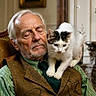 elderly_man, cat, indoor, closed_eyes, vintage_clothing, green_shirt, brown_vest, tie, pet, companion, shoulder, feline, white_cat, black_spots, relaxed, peaceful, home, portrait, close_up, warm_lighting