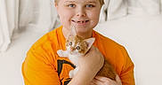 Cody joined the competition — help win amazing prizes! child, kitten, cat, orange_and_white, pet, hugging, portrait, smiling, indoors, bedroom, white_bedding, orange_shirt, short_hair, blue_eyes, animal, cute, cuddling, sitting, family, halloween