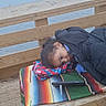 child, puffy_jacket, wooden_bench, colorful_blanket, smartphone, water, lake, outdoor, cloudy_sky, wooden_deck, casual, relaxed, portrait, side_view, young_person, winter_clothing, nature, daytime, calm, technology